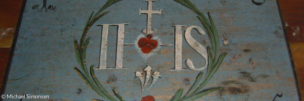 Ein bäuerlich-rustikales, himmelblau gestrichenes Holzbrett mit den Buchstaben IHS. In der Mitte ein flammendes Herz, darüber ein Kreuz, drum herum ein geschlossener Lorbeerkranz